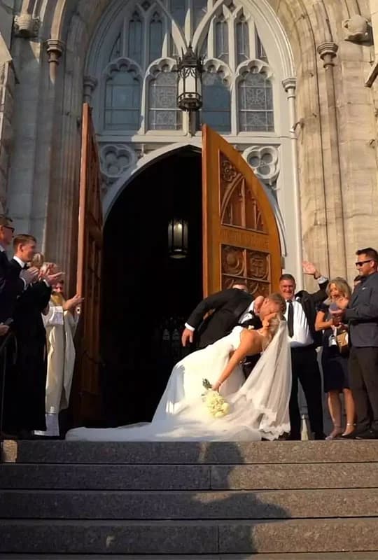 Caroline & Brennen in a dramatic dip kiss, standing on the steps of their church, being cheered on by their family and friends.