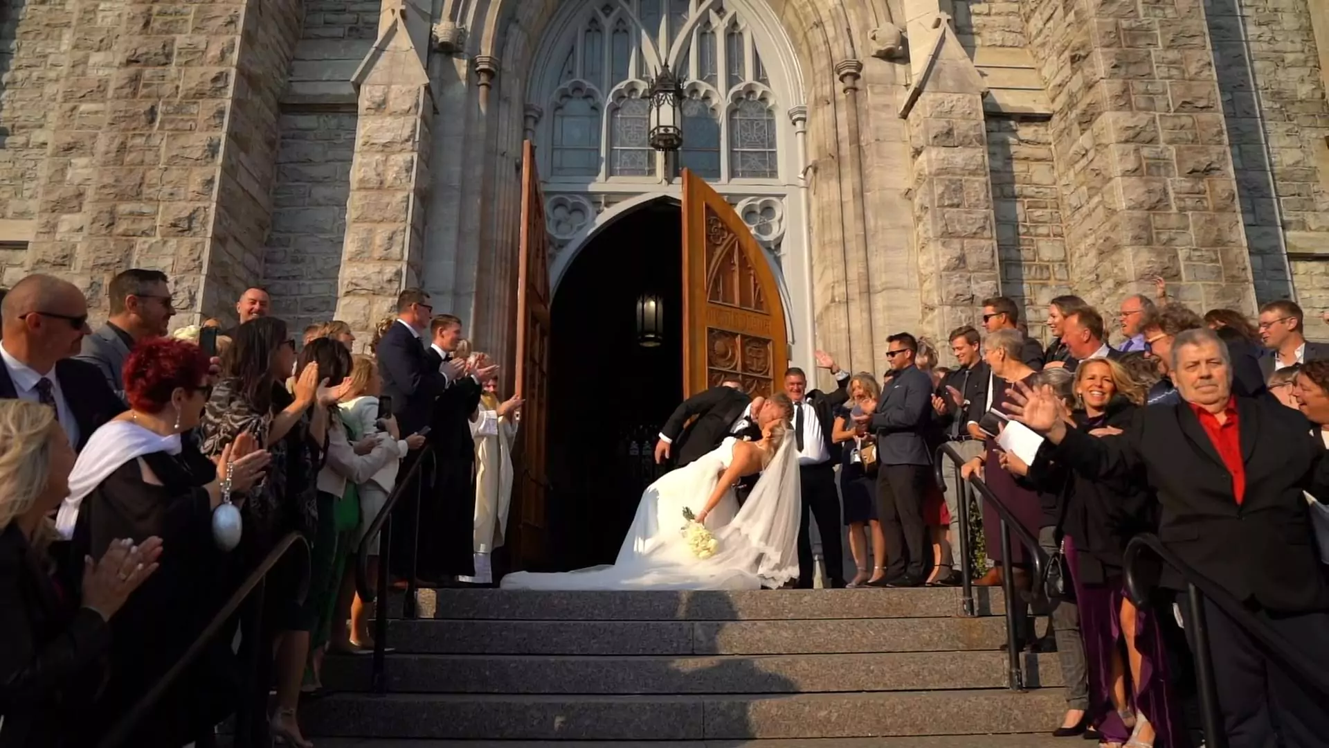 Caroline & Brennen in a dramatic dip kiss, standing on the steps of their church, being cheered on by their family and friends.
